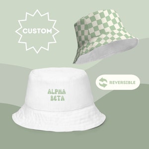 Puede incluir: Dos sombreros de pescador reversibles. Uno es blanco con "ALPHA BETA" en letras verdes. El otro sombrero tiene un patrón de tablero de ajedrez verde y blanco. La palabra "CUSTOM" está en forma de estrella. La palabra "REVERSIBLE" está al lado de un símbolo de flecha.