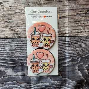 May include: Two round car coasters with a pink background and a design of two cartoon boba tea cups holding hands with a pink heart balloon above them. The text "Car Coasters" and "Handmade with love" is printed on the packaging.