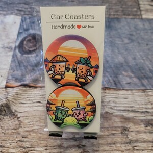 May include: Two sets of car coasters featuring a cartoon illustration of two boba tea cups with faces and arms, fighting. The background is a sunset with a pagoda. The text "Car Coasters" and "Handmade with love" is printed on the packaging.
