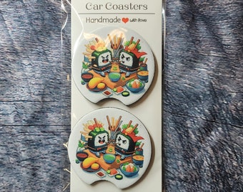 May include: Two round car coasters with a white background and a colorful design featuring two cartoon sushi rolls with chopsticks and other sushi ingredients. The text "Car Coasters" and "Handmade with love" is printed on the packaging.