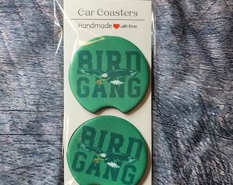 May include: Two green car coasters with the words "Bird Gang" and a green eagle graphic. The coasters are packaged in a clear plastic bag with a white card that says "Car Coasters Handmade with love".