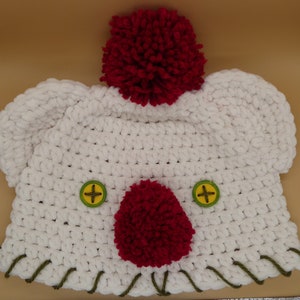 Puede incluir: Sombrero blanco de ganchillo con pompones rojos y botones verdes. El sombrero tiene un borde de ganchillo verde alrededor del borde inferior.