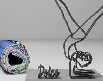 Peut inclure: Une sculpture en métal noir d'une personne faisant une pose de yoga, sur une base grise avec le mot "Dolce" écrit en noir. Un tapis de yoga roulé avec un motif coloré est en arrière-plan.