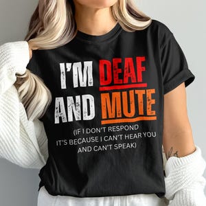 Peut inclure: Un t-shirt noir à manches courtes avec le texte blanc I'M AND, le texte rouge DEAF et le texte orange MUTE. En dessous, un texte blanc plus petit indique : IF I DON'T RESPOND IT'S BECAUSE I CAN'T HEAR YOU AND CAN'T SPEAK. Col rond.