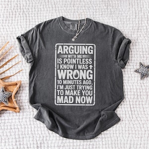 May include: A charcoal gray short-sleeve t-shirt featuring white text that reads: ARGUING WITH ME IS POINTLESS I KNOW I WAS WRONG 10 MINUTES AGO, I'M JUST TRYING TO MAKE YOU MAD NOW. The shirt has a crew neckline and a distressed, vintage-style graphic print.