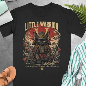 May include: A black t-shirt featuring a graphic of a black cat dressed in traditional samurai armor. The text LITTLE WARRIOR appears above the cat, with HONOR, COURAGE, CURIOSITY below. The background shows a red sun, cherry blossoms, and bamboo.