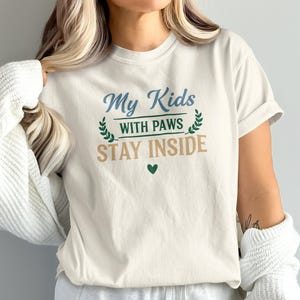 以下が含まれることがあります： 「My Kids with Paws Stay Inside」の文字が青、緑、茶色でプリントされたクリーム色のTシャツ。デザインにはハートと月桂樹の葉が含まれています。カジュアルで快適なスタイルです。