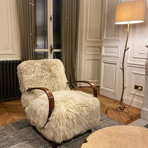 May include: A white faux fur armchair with wooden arms sits on a gray rug in a room with white walls and wooden floors. A gold floor lamp with a white shade is in the background.