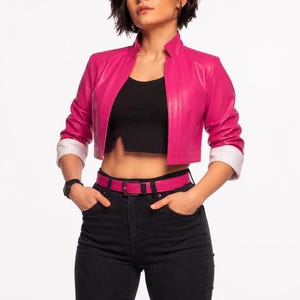 Op de afbeelding: Een korte, felroze leren jas met witte manchetten, gecombineerd met een zwarte top, zwarte jeans en een bijpassende roze riem. De outfit wordt gecompleteerd met een zwart horloge.