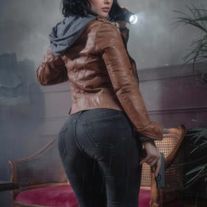 May include: A brown leather jacket with a gray hooded sweatshirt underneath, paired with black jeans. The person is holding a flashlight and a gun. The scene is set in a dimly lit, rainy environment.