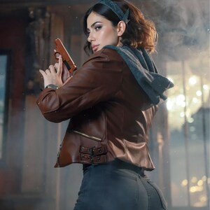 May include: A brown leather jacket with a gray hooded sweatshirt underneath. The jacket has a zipper closure and decorative buckles. The person is holding a brown handgun. The person is wearing dark gray jeans.