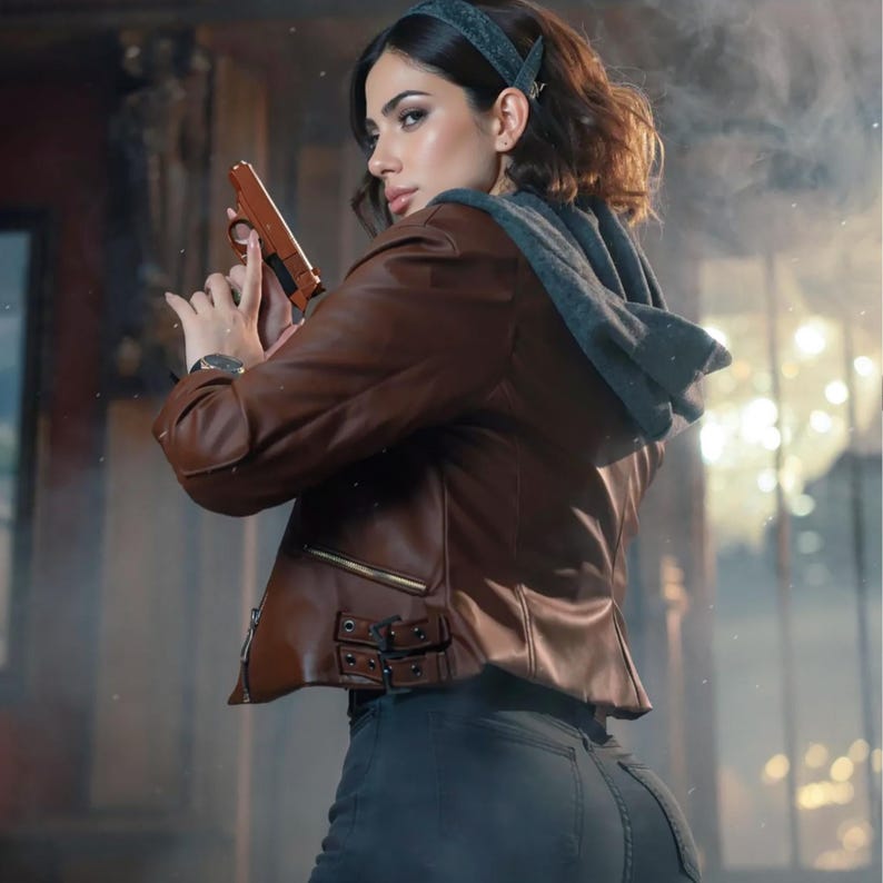 May include: A brown leather jacket with a gray hooded sweatshirt underneath. The jacket has a zipper closure and decorative buckles. The person is holding a brown handgun. The person is wearing dark gray jeans.