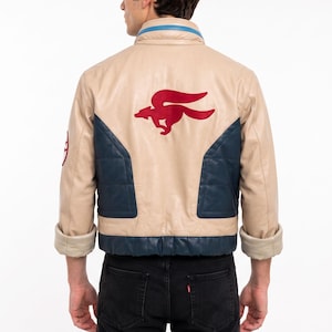 May include: A beige leather jacket featuring dark blue panels on the sides and lower back. A red winged fox emblem is centered on the back. The collar has a blue stripe, and a circular red patch is visible on the right sleeve. The cuffs are rolled up.