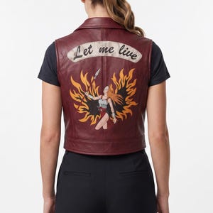 May include: A dark red leather vest featuring a back graphic of a figure holding a spear surrounded by orange and yellow flames. Above the graphic, a cream-colored banner displays the text "Let me live" in a black cursive script font.