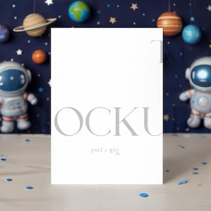 Könnte beinhalten: Eine weiße vertikale Karte mit dem Wort "OCKU" in grauen Buchstaben, darunter "psd + jpg". Die Karte befindet sich vor einem Weltraum-Hintergrund mit Planeten, Sternen und Astronautenfiguren. Blaue und goldene Luftballons befinden sich auf beiden Seiten.