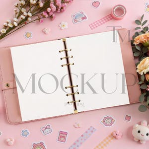 May include: A pink open planner with gold rings and blank pages, surrounded by stickers, flowers, and washi tape on a pink surface. The word "MOCKUP" is visible on the pages. The scene is a flat lay, ideal for stationery or craft product mockups.