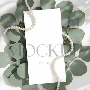 May include: A white rectangular card mockup with the word "MOCKUP" in gray, surrounded by green eucalyptus leaves and a pearl necklace. The card is centered on a white surface, with shadows.