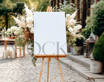 May include: A wooden easel with a blank white canvas stands on a stone path, adorned with floral arrangements of white roses, pampas grass, and greenery. The scene is set outdoors, suggesting a wedding or event.