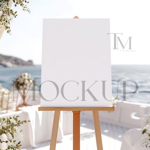 Puede incluir: Un cartel blanco en blanco sobre un caballete con un marco de madera. El cartel está colocado frente a una ceremonia de boda con flores blancas y sillas. El fondo es una vista borrosa del océano.