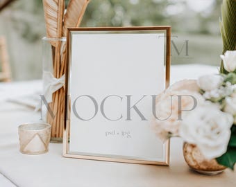 May include: A gold-framed mockup with the word "MOCKUP" in gray. The mockup is on a white surface, with a vase of dried plants and a small candle holder to the left, and a floral arrangement to the right.