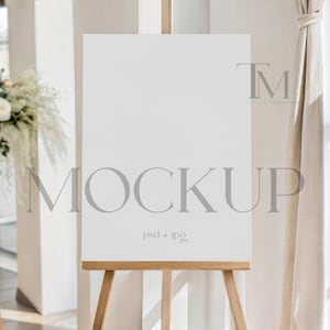 May include: A white rectangular sign on a wooden easel, with the word "MOCKUP" in gray letters. The sign is in a bright room with a floral arrangement and a window with natural light.