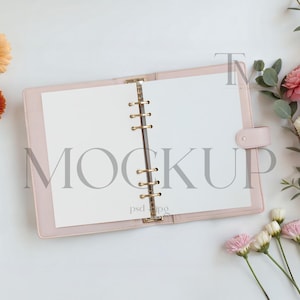 May include: A light pink planner with gold-colored rings and clasp, open to blank white pages. The word "MOCKUP" is printed in gray across the pages. The planner is surrounded by pink and orange flowers and greenery.