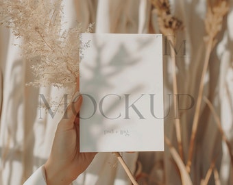 May include: A blank white card held in a hand against a backdrop of dried beige grasses and a blurred background. The card has the text "MOCKUP" and "psd + jpg" printed on it.