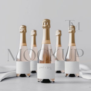 May include: Five champagne bottles with gold foil tops and blank white labels. The bottles are arranged on a light grey surface with a white cloth. The champagne inside the bottles has a light pink hue.