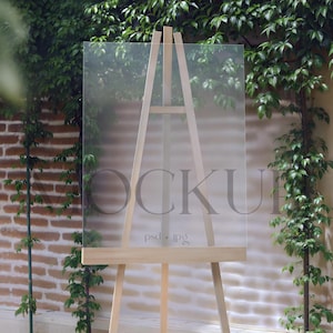May include: A clear acrylic sign on a wooden easel, with the word "MOCKUP" faintly visible. The background features a brick wall and lush green foliage, creating a natural setting. The text "psd.jpg" is also visible.