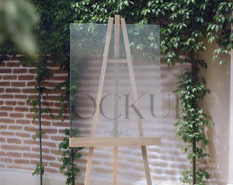 May include: A clear acrylic sign on a wooden easel, with the word "MOCKUP" faintly visible. The background features a brick wall and lush green foliage, creating a natural setting. The text "psd.jpg" is also visible.