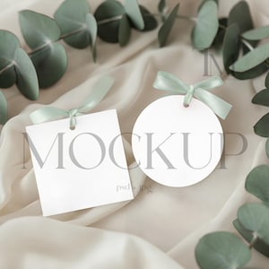 Pode incluir: Etiquetas brancas quadradas e redondas com fitas verde claro, dispostas sobre um tecido creme. A palavra "MOCKUP" está impressa nas etiquetas. Folhas de eucalipto circundam as etiquetas.