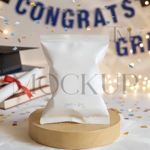 May include: A white snack bag stands on a wooden pedestal, with a graduation cap, books, and a diploma in the background. Confetti and string lights add to the celebratory atmosphere. The banner reads "CONGRATS GRAD".