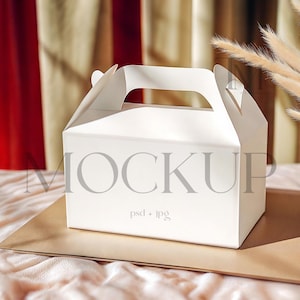 Puede incluir: Caja de cartón blanca con asa, ideal para empaquetar pequeños regalos o golosinas. La caja está sobre una superficie marrón con un fondo de tela suave de color rosa.