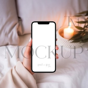 May include: A hand holding a black smartphone with a white screen. The phone is in a bed with white sheets and a lit candle in the background.