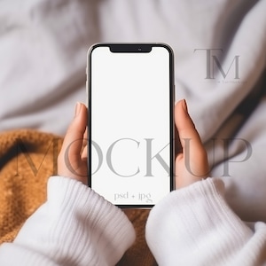 May include: A person is holding a white smartphone with a black frame. The screen is blank and the phone is in portrait mode. The person is wearing a white sweater and is sitting on a bed with a brown blanket.