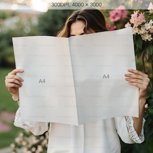 May include: A woman holds a blank sheet of paper in front of her face. The paper is folded in half and has the text "A4" printed on both sides. The woman is standing in front of a bush of pink flowers.