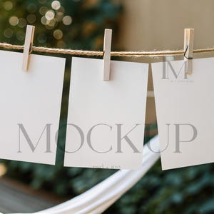 May include: Three blank white cards are hanging on a clothesline with wooden clothespins. The cards are in a row and are all the same size. The background is blurry and out of focus, but it appears to be a garden or outdoor setting.