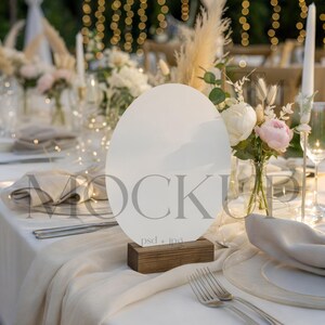 May include: A wedding table setting featuring a white oval sign on a wooden base, with the word "MOCKUP" printed on it. The table is set with white linens, silverware, and floral arrangements. The background includes string lights and a blurred outdoor scene.