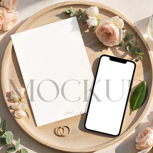 Puede incluir: Una hoja de papel rectangular blanca en blanco y un teléfono inteligente con una pantalla blanca en blanco colocados sobre una bandeja redonda de madera. La bandeja está decorada con rosas rosa pálido, hojas de eucalipto verdes, dos anillos dorados y un vaso de agua con limón.