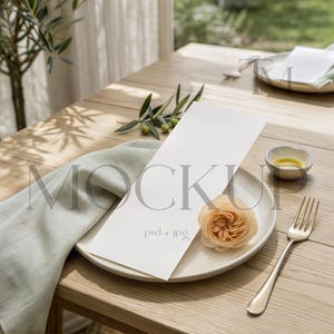May include: A blank white rectangular menu card placed on a round ceramic plate, accompanied by a light green linen napkin, a peach-coloured flower, a silver fork, a small bowl with yellow liquid, and a clear glass on a wooden table.