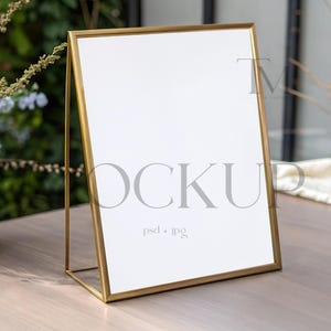 May include: A gold-colored easel with a blank white sign. The easel is on a wooden table with a vase of flowers in the background.