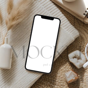 May include: A smartphone with a blank white screen rests on a cream-colored knitted sweater. A white ceramic vase with dried pampas grass, a cup of tea, and decorative crystals are also present. A notebook and woven mat complete the scene.