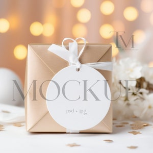 May include: A gold gift box with a white ribbon and a white round tag that says "MOCKUP psd + jpg" on a white surface with gold confetti and a blurred background of white lights.