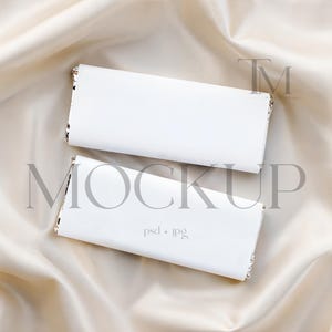 Puede incluir: Dos objetos rectangulares blancos con ribete plateado están dispuestos sobre una tela de satén color crema. El objeto inferior tiene el texto "psd + jpg" impreso. Probablemente sean maquetas de productos.