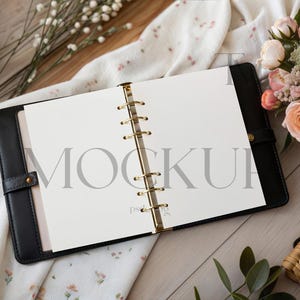 May include: Black leather binder with gold-coloured rings, open to reveal blank white pages. The word "MOCKUP" is printed in light grey across the pages. The binder is on a wooden surface, with flowers and fabric in the background.