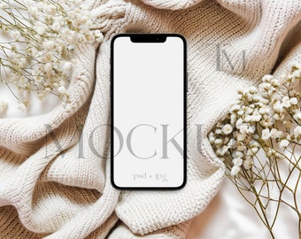 May include: A black smartphone with a blank screen rests on a cream-colored knitted blanket, surrounded by white baby's breath flowers. The words "MOCKUP" and "psd + jpg" are visible.