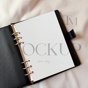 May include: Black leather binder with gold-colored rings, open to reveal a blank white page. The word "MOCKUP" is printed in gray across the page. The binder is on a light-colored fabric background.