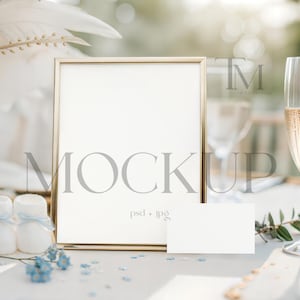 May include: A gold framed mockup with a blank white space, a small white card, and a champagne flute filled with a bubbly beverage. The scene is decorated with white flowers, candles, and blue accents.