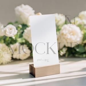 Op de afbeelding: Een witte rechthoekige kaart in een houten houder, met het woord "MOCKUP" in grijze letters. De kaart staat voor een wazige achtergrond van witte bloemen en groen. De tekst "psd + jpg" staat onder het woord "MOCKUP".