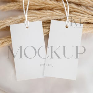以下が含まれることがあります： 白い長方形の空白のタグが2つ、ひもで吊り下げられており、乾燥したベージュ色のパンパスグラスと白いサテン生地を背景にしています。「MOCKUP」の文字がグレーでタグに印刷されています。「psd + jpg」の文字が下に印刷されています。